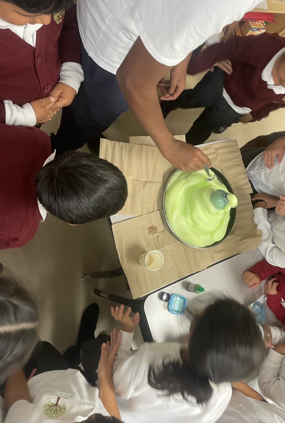 Students crowded around a tray watching a green foam reaction overflow a bottle