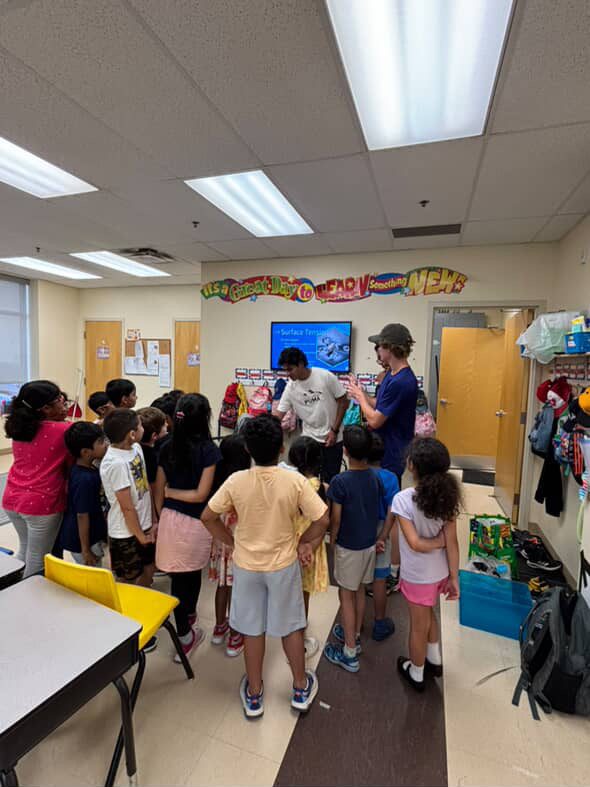 Students gathered around two STEMroots instructors in a classroom during a surface tension lesson