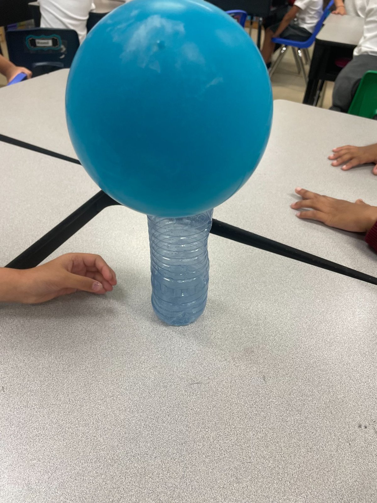 A balloon inflating on top of a bottle during the baking soda and vinegar reaction, with students’ hands on the table